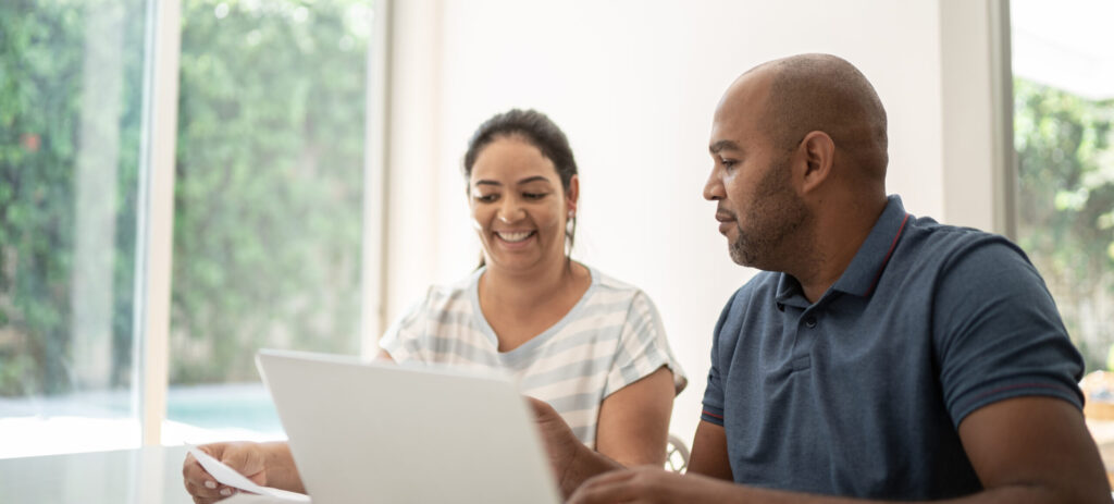 A woman and man sit next to each other at a table looking at some papers and a computer screen.